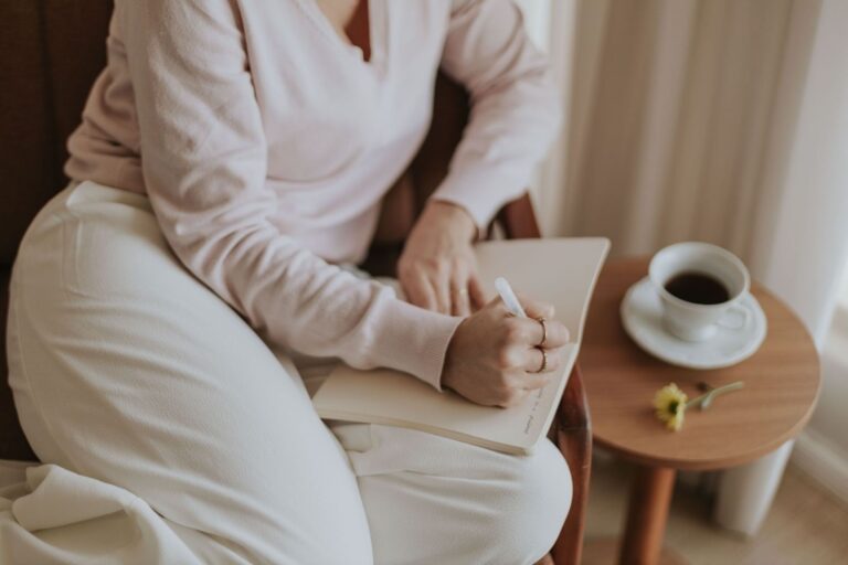 Person i ljusa kläder skriver i en anteckningsbok med kaffe bredvid